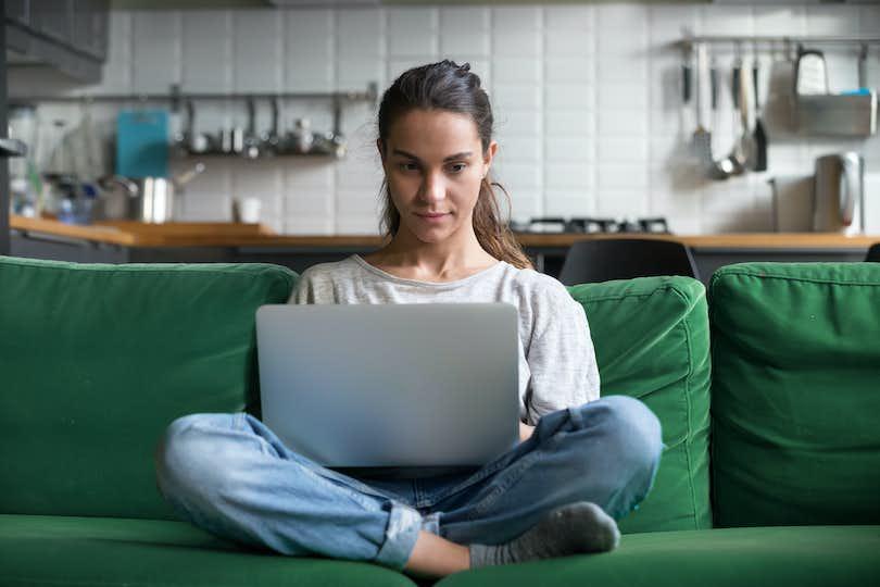 a woman sits cross-legged on a sofa staring intently at the laptop situated on her lap